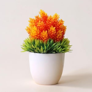 May include: A small, artificial plant in a white ceramic pot. The plant features vibrant orange and yellow flower-like structures atop a bed of green, grass-like foliage. The pot is smooth and rounded, creating a simple, modern aesthetic.