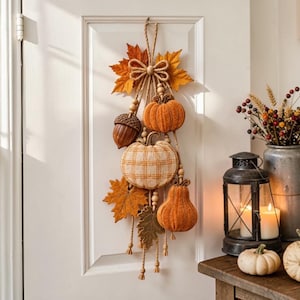 Op de afbeelding: Een hangende herfstwanddecoratie met stoffen pompoenen, een grote eikel en kunstmatige esdoornbladeren in tinten oranje, geel en bruin. De elementen zijn opgehangen aan jutekoord met een decoratieve strik en houten kralen.