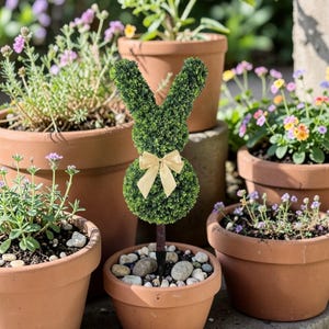 Könnte beinhalten: Ein Buchsbaumkaninchen mit goldener Schleife, in einem Terrakottatopf mit Kieselsteinen. Das Kaninchen ist von anderen Topfpflanzen mit bunten Blumen umgeben, wodurch eine Gartenszene entsteht.