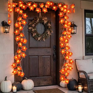 Puede incluir: Una puerta de madera oscura decorada con una corona de hojas marrones secas y una guirnalda de luces de hojas de arce naranjas alrededor del marco. Varias calabazas blancas y negras están en el suelo con velas encendidas dentro de farolillos.