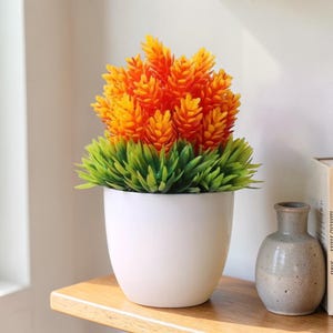 May include: A white ceramic pot filled with a vibrant artificial plant. The plant features bright orange and yellow flower clusters atop a bed of green, spiky leaves. The pot sits on a wooden shelf, with a small gray vase and books in the background.