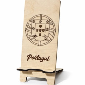 Peut inclure: Support de téléphone en bois clair avec les armoiries portugaises et le mot "Portugal" en écriture cursive. Le support a une fente pour tenir un téléphone et une base pour la stabilité.