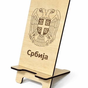 May include: A wooden phone stand with a laser-etched design of the Serbian coat of arms, featuring a double-headed eagle and the word "Србија". The stand is made of light-colored wood and has a slot to hold a phone.