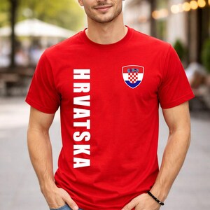May include: Red t-shirt with the word "HRVATSKA" in white vertical text on the left side. A Croatian coat of arms is on the right chest. The shirt is worn with blue jeans.