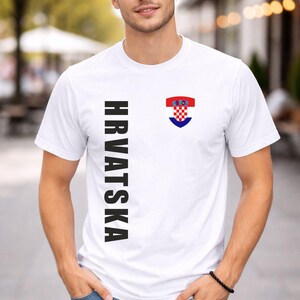 May include: White t-shirt with the word "HRVATSKA" printed vertically in black on the left side. A Croatian coat of arms is on the right chest. The shirt is short-sleeved.