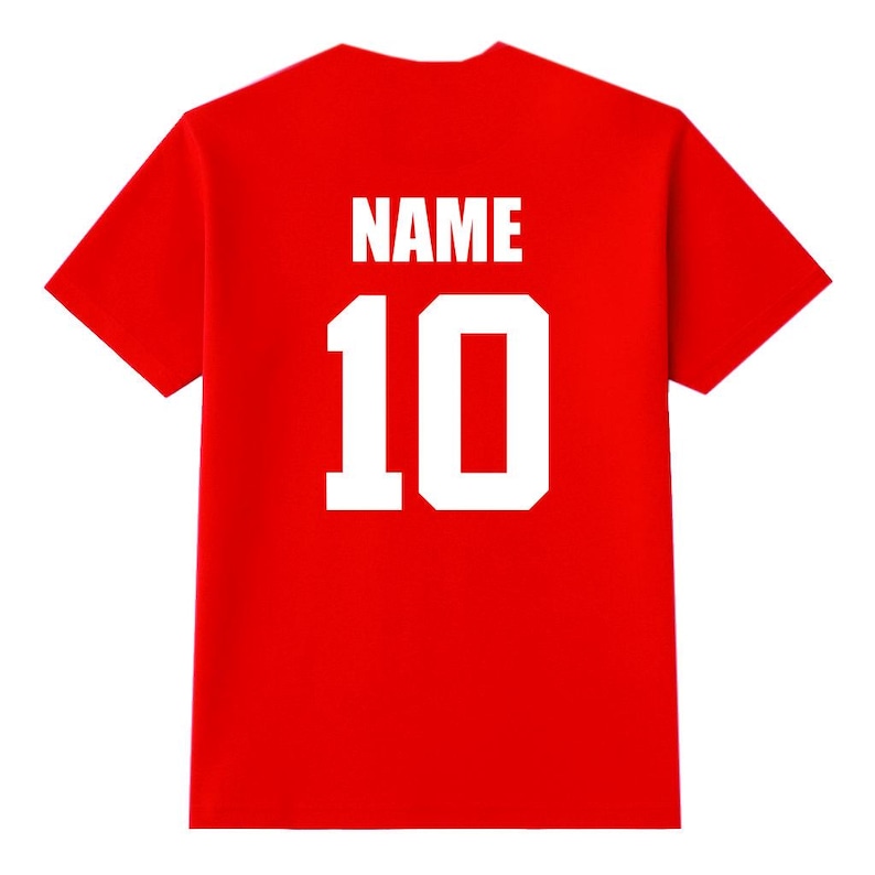 May include: A red t-shirt with the word "NAME" and the number "10" printed in white block letters on the back. The shirt is a solid red color and has short sleeves.