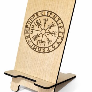 Puede incluir: Soporte de teléfono de madera con un símbolo Vegvísir grabado con láser. El soporte está hecho de madera de color claro con un diseño circular marrón oscuro. El soporte está inclinado para facilitar la visualización y tiene una ranura para sostener un teléfono.