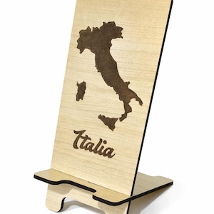 May include: A wooden phone stand featuring a laser-etched map of Italy. The word "Italia" is written in a script font below the map. The stand is made of light-colored wood with a dark brown outline of the country.