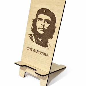 Peut inclure: Un support de téléphone en bois clair avec un portrait de Che Guevara et le texte "CHE GUEVARA" en marron. Le support a un design simple et fonctionnel, idéal pour un bureau.