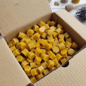 May include: A cardboard box filled with numerous yellow, hexagonal wax cubes. The cubes are a warm, golden color, suggesting a natural beeswax product. The box is open, revealing the contents.