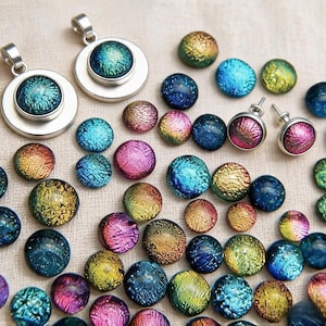 May include: A collection of circular dichroic glass cabochons in iridescent shades of blue, green, gold, and pink, alongside two silver-toned metal pendants and a pair of matching silver-toned stud earrings featuring the same glass design.