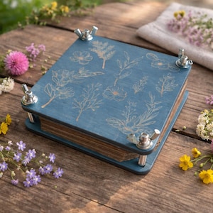 May include: A blue wooden flower press with botanical illustrations and wing nuts. The press is surrounded by colorful wildflowers on a rustic wooden surface. A linen cloth with flowers is in the background.