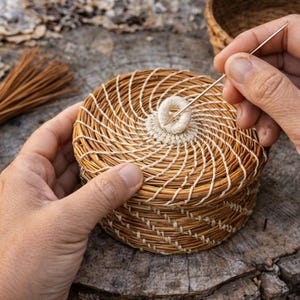 May include: A close-up of a small, round, woven basket with a lid. The basket is made of natural-colored fibers and is being sewn with a needle and thread. A bundle of brown fibers and a larger basket are in the background.