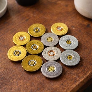 May include: A collection of metallic shotgun shell casings arranged on a wooden surface. The casings are in shades of gold and silver, with the text "12 GA" visible on some. The arrangement suggests a decorative or repurposed item.