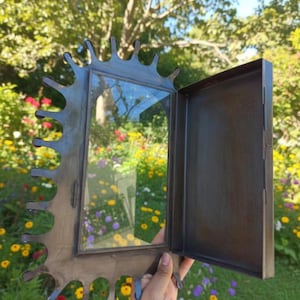 May include: A decorative metal frame with a hinged door, featuring a unique, irregular edge design. The frame has a dark, weathered finish and a clear glass panel. The door is open, revealing the interior. The frame is held against a backdrop of colorful flowers.