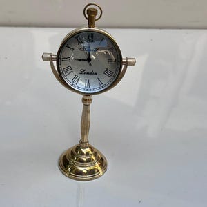 May include: A gold-toned metal desk clock with a circular face, Roman numerals, and a pedestal base. The clock face features the text Smith and London. It has a small ring at the top and a swivel mechanism connecting the clock to the stand.