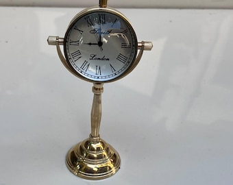 Op de afbeelding: Een goudkleurige metalen bureauklok met een ronde wijzerplaat, Romeinse cijfers en een voetstuk. Op de wijzerplaat staat de tekst Smith en London. De klok heeft een ring aan de bovenkant en een draaimechanisme op de standaard.
