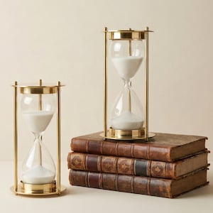 Könnte beinhalten: Zwei Sanduhren mit goldfarbenen Metallrahmen und durchsichtigen Glaskolben, die mit weißem Sand gefüllt sind. Eine Sanduhr steht allein, während die andere auf einem Stapel von drei antiken Büchern mit braunen Lederdeckeln ruht.