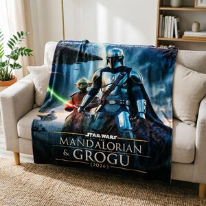 Pode incluir: Uma manta de pel&uacute;cia com um design de Star Wars com The Mandalorian e Grogu. A manta tem um esquema de cores azul e cinza com o texto "STAR WARS THE MANDALORIAN & GROGU (2026)". Est&aacute; sobre um sof&aacute; bege.