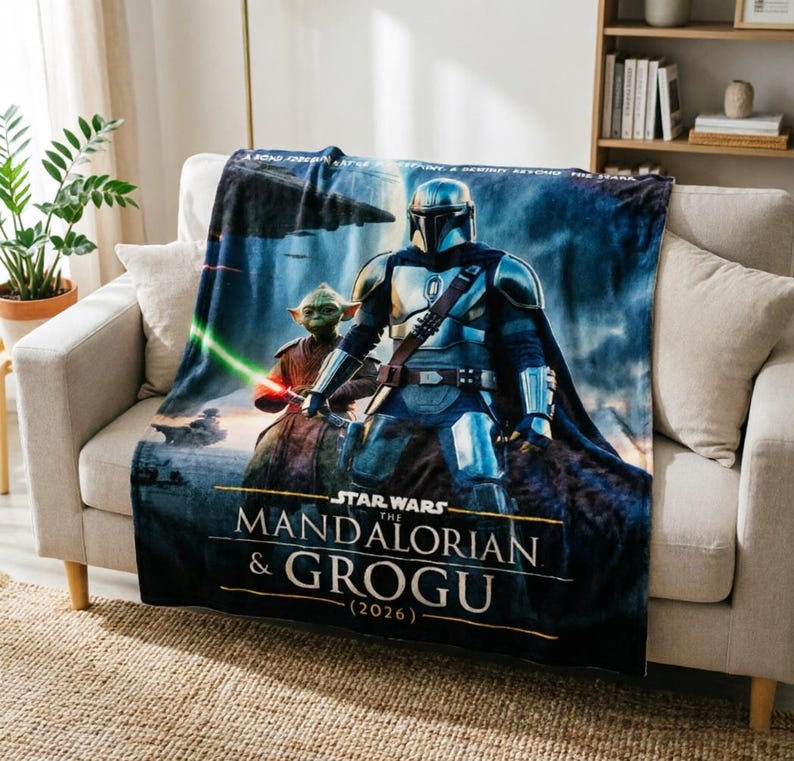 Pode incluir: Uma manta de pel&uacute;cia com um design de Star Wars com The Mandalorian e Grogu. A manta tem um esquema de cores azul e cinza com o texto "STAR WARS THE MANDALORIAN & GROGU (2026)". Est&aacute; sobre um sof&aacute; bege.
