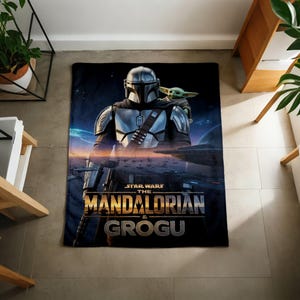 Pode incluir: Um cobertor preto com o Mandaloriano e Grogu de Star Wars. O Mandaloriano est&aacute; em armadura prateada, e Grogu est&aacute; em seu ombro. O cobertor tem as palavras "STAR WARS THE MANDALORIAN & GROGU" em letras douradas.