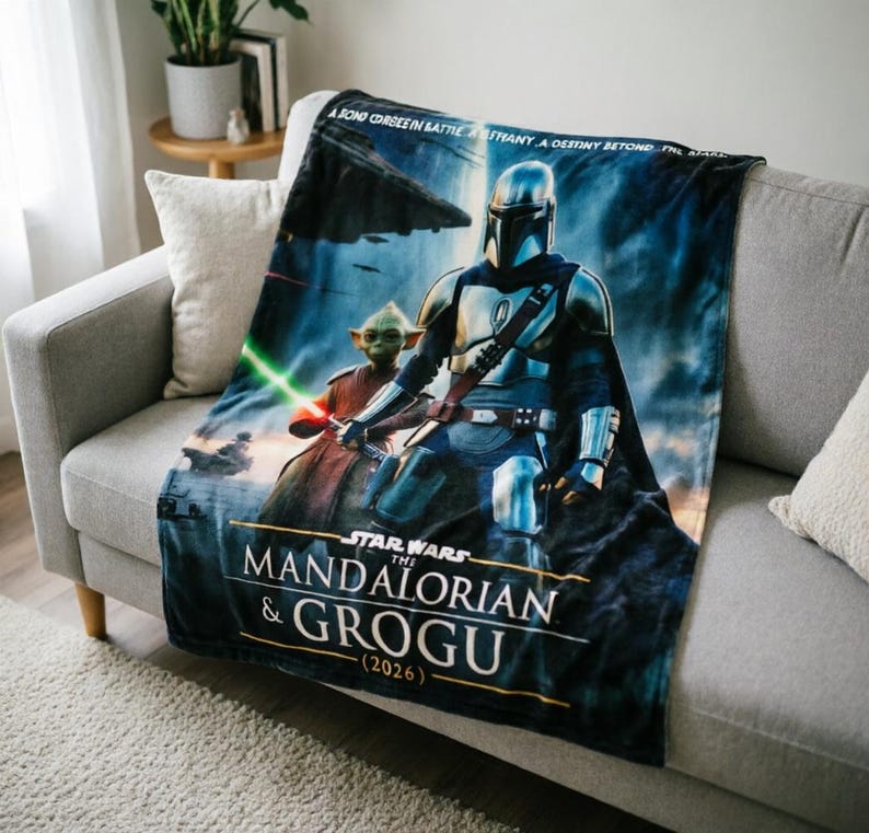 Pode incluir: Um cobertor com o tema Star Wars com The Mandalorian e Grogu. O cobertor tem um fundo azul com imagens dos personagens e o texto "The Mandalorian & Grogu (2026)". O cobertor est&aacute; sobre um sof&aacute; cinza.