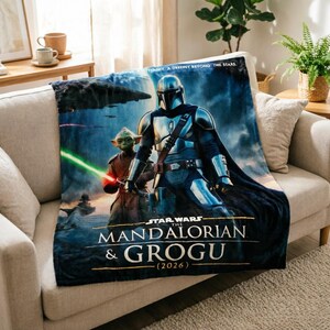 Pode incluir: Uma manta de pel&uacute;cia com personagens de "The Mandalorian". O design inclui o Mandaloriano e Grogu, com o texto "STAR WARS THE MANDALORIAN & GROGU (2026)". A manta est&aacute; sobre um sof&aacute; bege.