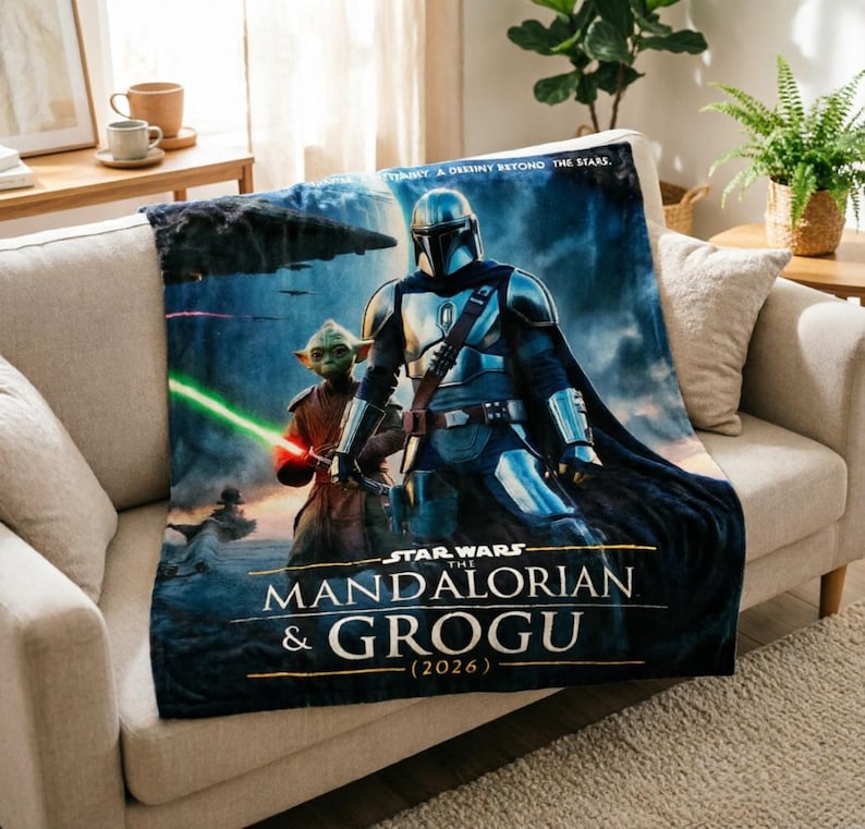 Pode incluir: Uma manta de pel&uacute;cia com personagens de "The Mandalorian". O design inclui o Mandaloriano e Grogu, com o texto "STAR WARS THE MANDALORIAN & GROGU (2026)". A manta est&aacute; sobre um sof&aacute; bege.