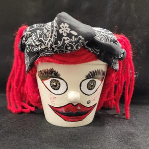 May include: A hand-painted, off-white ceramic flower pot decorated to resemble a face. It features red yarn hair, large painted eyes, red lips, and a black and white bandana. A whimsical, decorative item.
