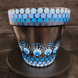 May include: A black plant pot featuring a hand-painted dot art design in shades of light blue, medium blue, and dark blue. The rim displays horizontal rows of dots, while the body showcases a scalloped pattern of concentric circles and dots.