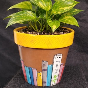 Puede incluir: Una planta en maceta con hojas verdes y amarillas en una maceta de terracota marrón. La maceta tiene un borde amarillo y está decorada con libros pintados de colores y la palabra "Plants".