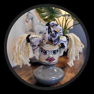 May include: A whimsical ceramic vase shaped like a woman's head, with painted facial features and yarn hair. The head is adorned with a patterned scarf and black studded accents. The vase is gray and sits on a wooden surface.