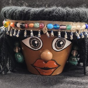 Peut inclure: Pot de fleur en terre cuite décoré pour ressembler à un visage. Il présente de grands yeux peints, des lèvres rouges et un bandeau de perles. De la laine noire forme les cheveux, et des boucles d'oreilles pendantes complètent le look. Une pièce artistique unique.