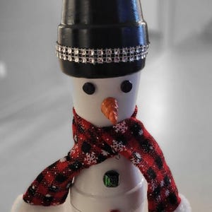 Könnte beinhalten: Handgefertigte Schneemann-Dekoration aus bemalten Töpfen. Er hat einen schwarzen Zylinder mit Strassband, einen rot-schwarz karierten Schal und dekorative Knöpfe. Der Schneemann hat eine Karottennase und einen Kristall-Edelstein.