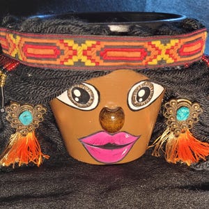 Puede incluir: Una maceta decorativa con forma de rostro con una cara marrón, ojos grandes y labios rosas. Presenta cabello de hilo negro, una diadema estampada y pendientes ornamentados con piedras turquesas y borlas naranjas. La maceta está diseñada para contener una planta pequeña.
