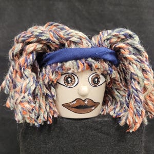 May include: A small, light-colored ceramic pot painted with a face featuring brown eyes and dark lips. The pot is adorned with a thick, textured mop of multi-colored yarn in shades of blue, orange, and cream, secured by a solid dark blue fabric headband.