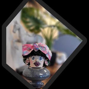 Pode incluir: Um vaso de flores decorativo pintado à mão em forma de rosto feminino. O vaso apresenta um lenço rosa e azul estampado, cabelo preto, olhos azuis grandes e brincos rosa. Ele está em cima de um vaso cinza bulboso.