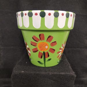 May include: A lime green terracotta plant pot featuring hand-painted orange flowers with dark brown petals. The rim is decorated with a repeating pattern of white vertical stripes, dark green circles, and small red dots against a white background.