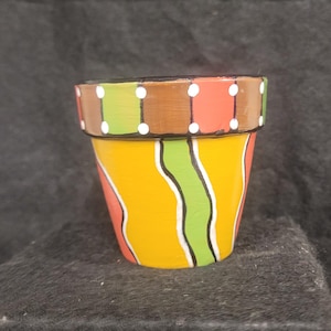 May include: A small, hand-painted ceramic flower pot. The pot is painted with a vibrant yellow base, accented by vertical green and red stripes. The rim features a brown band with white polka dots. A unique and colorful decorative item.