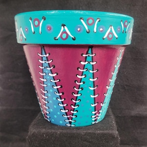Op de afbeelding: Een handbeschilderde terracotta plantenpot met een levendige turquoise rand met paarse en witte cirkelvormige patronen. De romp toont afwisselende verticale panelen van diep magenta en turquoise, versierd met witte stikselachtige lijnen.