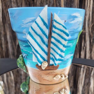 May include: A hand-painted flower pot with a nautical theme. The pot features a blue sky and ocean scene with a sailboat made of wood and fabric. Seashells are glued to the base. The pot is a light brown color.