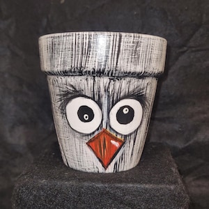 Op de afbeelding: Handbeschilderde keramische bloempot met een grillig vogelgezichtontwerp. De pot heeft grote witte ogen met zwarte pupillen, een rood-oranje snavel en een getextureerde grijze en witte afwerking. Ideaal voor binnen of buiten.