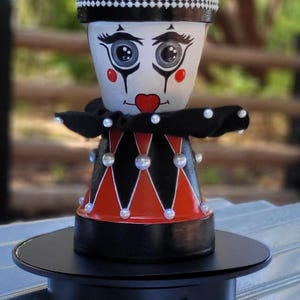 Pode incluir: Um vaso de flores pintado à mão com tema de palhaço, com uma borda xadrez em preto e branco, um rosto pintado e uma gola preta adornada com pérolas. O vaso tem um padrão de losangos vermelhos e pretos e é preenchido com vegetação. Ele está em uma base giratória preta.