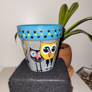 Könnte beinhalten: Handbemalter grauer Keramik-Blumentopf mit drei Cartoon-Eulen. Der Topf hat einen blauen Rand mit bunten Punkten. Die Eulen sind gelb und schwarz mit großen Augen. Der Topf steht auf einer dunkelgrauen Oberfläche.