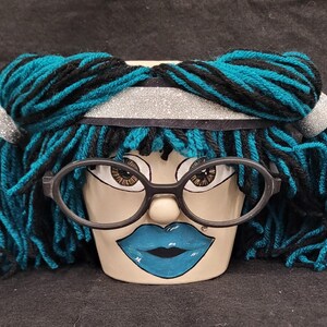 May include: A ceramic planter shaped like a face, featuring thick teal and black yarn hair styled in pigtails with a silver glitter headband. The face has painted brown eyes, a small nose, and bright blue lips, accessorized with black glasses.