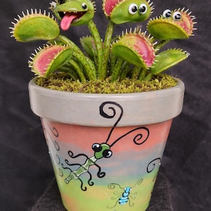 Puede incluir: Una planta carnívora Venus pintada a mano en una maceta decorativa. Las trampas verdes y rosas tienen caras de dibujos animados, y una mosca está posada en una de ellas. La maceta está pintada con un diseño colorido de insectos y espirales.