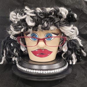 May include: A ceramic planter shaped like a face with painted blue eyes, long eyelashes, and red lips. It wears red-rimmed glasses, dangling clear crystal-like earrings, and a thick, curly black and white yarn wig. The base is black with rhinestones.