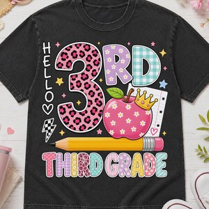May include: A black t-shirt featuring a graphic with the text HELLO 3RD THIRD GRADE. The design includes a large number 3 with a pink leopard print, a pink apple with white flowers and a gold crown, a yellow pencil, and colorful patterned letters.