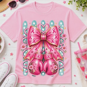 May include: A light pink t-shirt featuring a graphic print of two pink boxing gloves adorned with floral patterns and a large pink bow. The bow is decorated with small pink breast cancer awareness ribbons and hearts, set against a teal polka dot background.