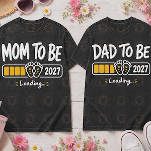 May include: Two black t-shirts featuring white text and graphics. The left shirt reads MOM TO BE, and the right reads DAD TO BE. Both include a yellow loading bar, baby footprints, the year 2027, and the text Loading... in white and yellow.